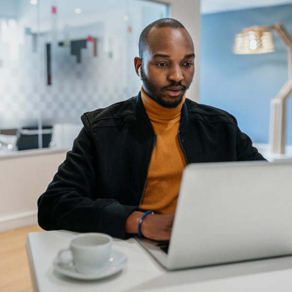 Focused black African American millennial man sitting at desk using laptop and earpods in coworking space