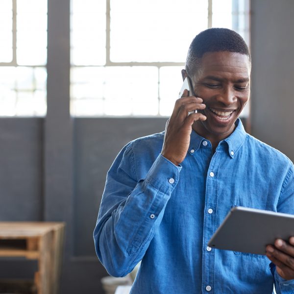 Smiling young African businessman standing in a large modern office working on a digital tablet and talking on a cellphone