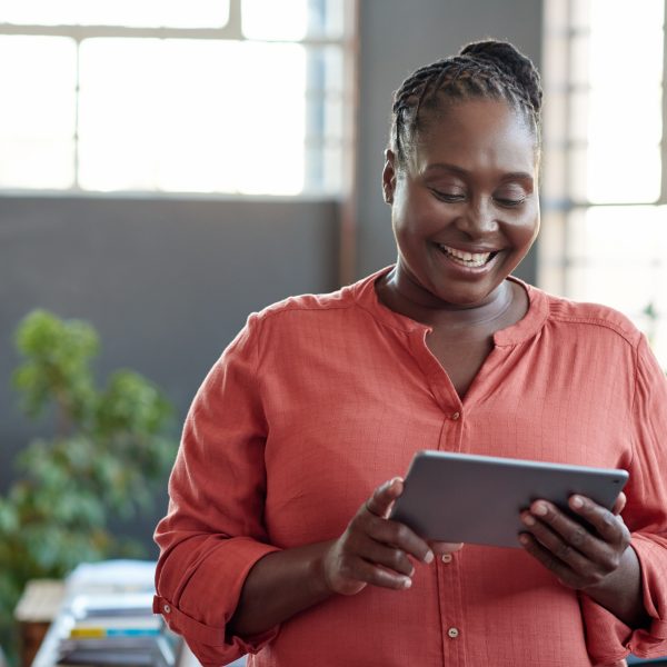 Casually dressed young African businesswoman smiling and using a digital tablet while standing alone in a modern office